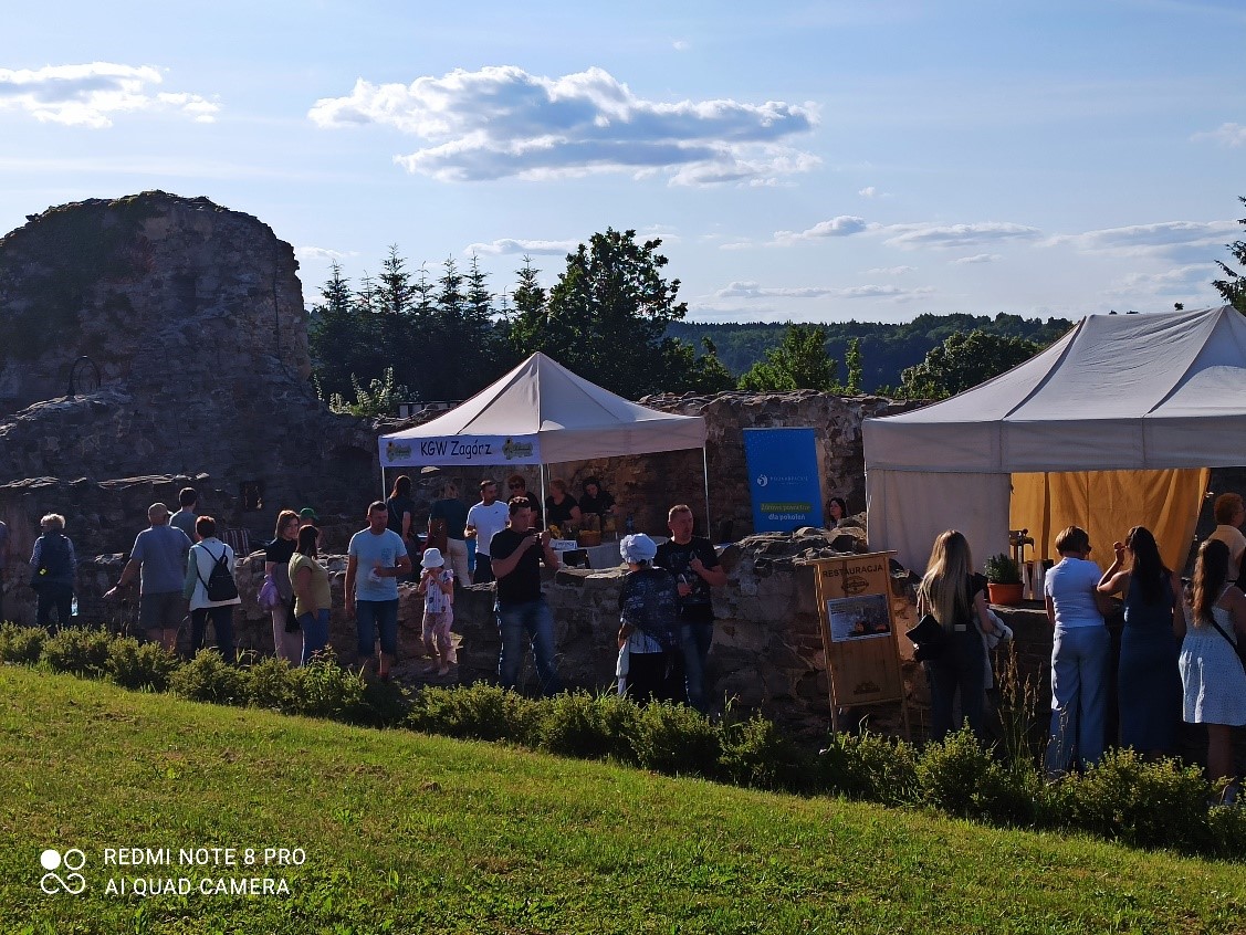 Targ na świeżym powietrzu. W tle ruiny zamku i stoiska handlowe pod namiotami, otoczone przez spacerujących ludzi.